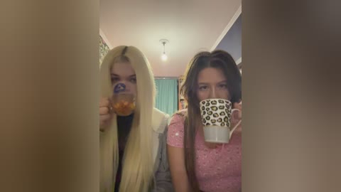 Video of two young women, one with long blonde hair and another with dark hair, holding cups, standing in a hallway with teal curtains and a ceiling light.
