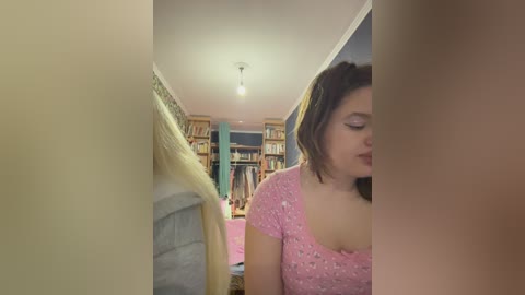 Video of a woman with short brown hair in a pink floral top, sitting in a cozy, dimly lit room with bookshelves and a bed, partially obscured by a white curtain.