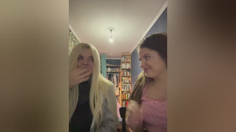 Video of two teenage girls with long hair in a narrow hallway, one hiding her face, the other smiling, surrounded by bookshelves.