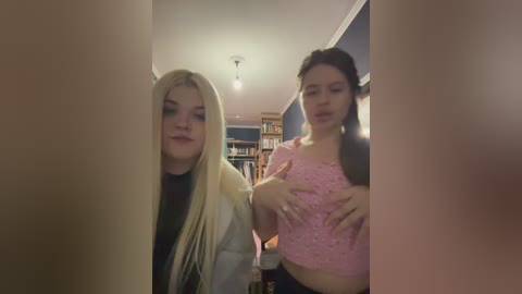 Video of two young women, one with long blonde hair, wearing a white jacket, the other with brown hair, in a pink top, standing in a dimly lit room with bookshelves and a ceiling light.