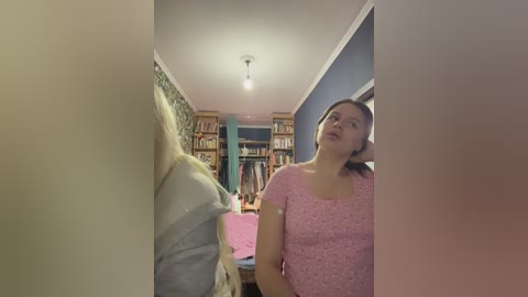Video of a woman with brown hair in a pink floral dress standing in a hallway, looking at a white dress hanging on a door. Background includes shelves of books and a pink table.