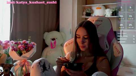 Media: A video of a young woman with long brown hair, wearing a black top, sitting in a pink gaming chair, eating with a spoon, in a cozy, floral-decorated room.