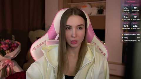 Media: Video of a young woman with straight brown hair wearing a pink and white gaming headset, sitting in a cozy room with dim lighting.