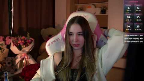 Media: Video of a young woman with long brown hair, wearing a white hoodie, sitting in a cozy room with a large teddy bear and a floral arrangement in the background.