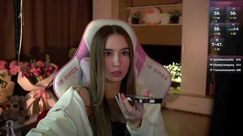 Media: A video of a young woman with long blonde hair in a pink gaming chair, holding a smartphone, in a dimly lit room with shelves and flowers in the background.