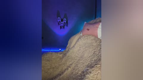 Video of a plush dog toy on a beige blanket, illuminated by a blue light. The toy, with a pink paw and white muzzle, sits in a dimly lit room with a shadowy wall featuring a cartoon dog.
