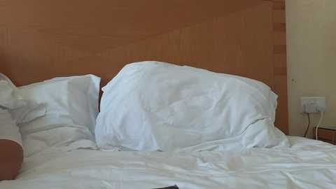 Video of a messy hotel bed with rumpled white sheets and pillows against a light wooden headboard. A white electrical outlet and a small white appliance are visible on the right wall.
