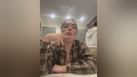 Video of a young woman with light skin, dark hair, and a plaid shirt, blowing smoke from a cigarette, in a dimly lit bathroom with white tiles and a towel.