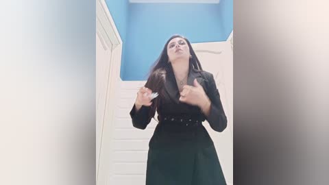 Media: Video of a woman with long dark hair, wearing a black blazer and skirt, standing in a narrow, white-paneled hallway with blue walls. She's in a defensive pose, hands raised.