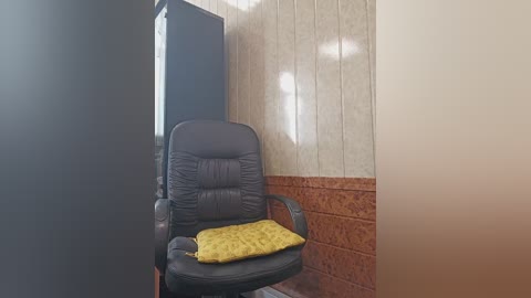 Media: Video of a dark leather office chair with a yellow cushion, situated in a room with beige and brown tiled walls, and a black wall on the left.