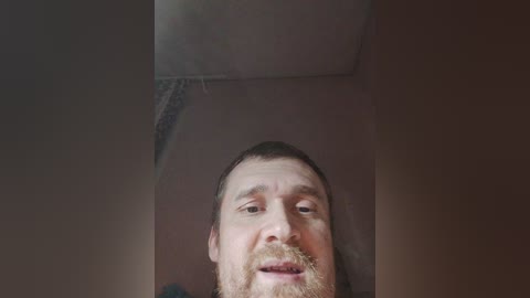 Media: A close-up video of a bearded Caucasian man with light skin and short brown hair, looking up with a neutral expression. The background is a dimly lit, beige ceiling.