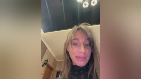 A video of a young woman with light skin and long, straight, blonde hair. She has a slender build and wears a black scarf. The background features a modern ceiling with recessed lights and a brick wall.