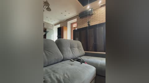 A video of a cozy, partially lit living room featuring a light gray sectional sofa, wooden blinds, and a brick wall, with a ceiling fan and an air conditioner visible.