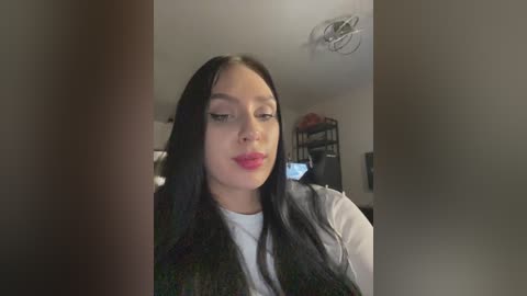 Media: Video of a young woman with long black hair, wearing a white top and dark vest, standing in a dimly lit room with shelves and a TV in the background.