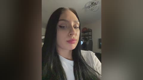 Media: Video of a young woman with long, straight black hair, fair skin, and light pink lipstick, wearing a white t-shirt and dark vest, indoors with blurred background of shelves and a ceiling fan.