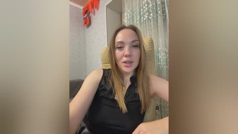 Video of a Caucasian woman with light skin and straight brown hair, wearing a black sleeveless top, seated in a beige armchair with a festive orange ribbon and a pumpkin decoration on the wall.