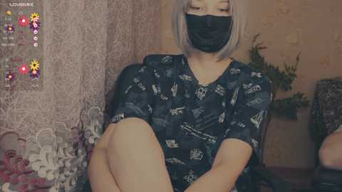 A video of a young woman with short, platinum blonde hair, wearing a black face mask, black top with white patterns, and sitting on a bed with floral bedding, in a cozy, dimly lit room.