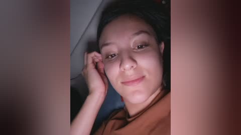 A video of a smiling woman with medium brown skin, dark hair, and brown eyes, resting her head on her hand, wearing a brown jacket, taken from a close-up perspective.
