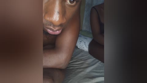Video of a dark-skinned, shirtless man lying on a bed, partially obscured by a person's arm, with a blurred, dimly lit background.