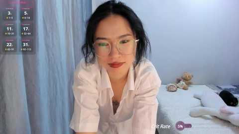 Video of an Asian woman with fair skin, wearing glasses, a white blouse, and a necklace, leaning forward on a bed in a softly lit room.