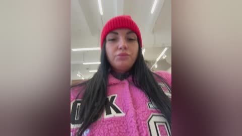 A video of a young woman with long black hair, wearing a red beanie and a pink hoodie with large white numbers, standing indoors under bright overhead lights.