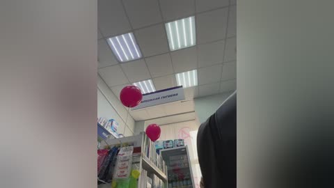 A video of a brightly lit, modern pharmacy with fluorescent lights, white ceiling tiles, and pink balloons hanging from a sign above shelves filled with products.