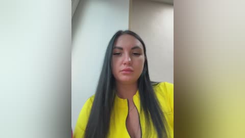 Video of a woman with long, straight black hair and medium skin tone, wearing a bright yellow shirt, standing in a dimly lit room with white walls.