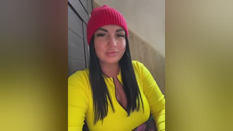 Video of a woman with long black hair, wearing a bright yellow long-sleeve top and a red knit beanie, standing indoors near a wooden wall.