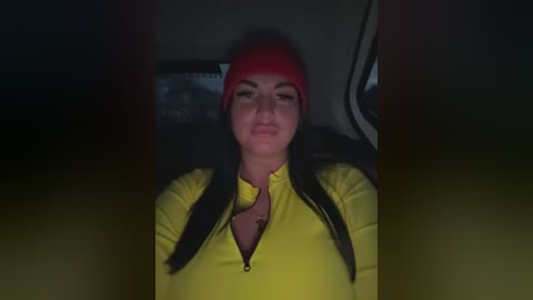 Video of a woman with medium-length black hair, wearing a bright yellow jacket, red beanie, and a necklace. She sits in a dimly lit car with a dark background.
