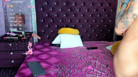 Video of a bed with a purple tufted headboard, a pink bra on the floor, a laptop, and a TV remote on a pink floral bedspread.