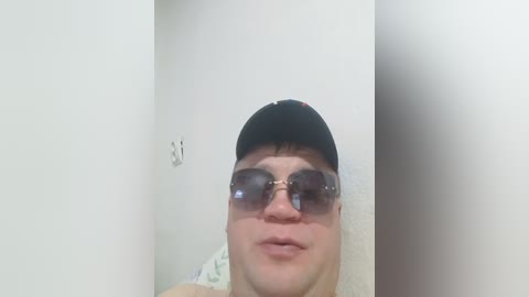 Video of a young, fair-skinned person with short, dark hair, wearing sunglasses, a black cap, and a light-colored shirt, standing in a dimly lit hallway with white walls and a patterned blanket visible.