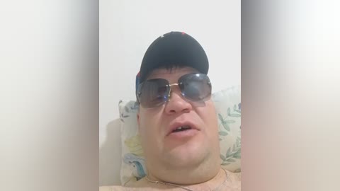Video of a Caucasian man with glasses, a black cap, and a floral pillow in a white room. He has a slightly open mouth and a neutral expression.