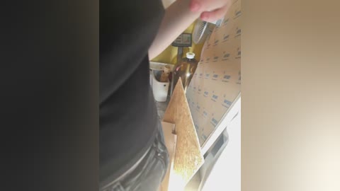 Media: Video of a person in a black shirt, holding a bottle, pouring liquid onto a wooden board covered in blue stickers, in a kitchen with yellow walls and a white counter.