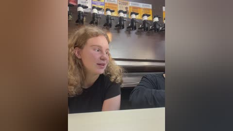 Media: Video of a young woman with curly blonde hair, wearing a black t-shirt, sitting at a bar counter. Behind her, black and white cowboy boots hang on a wall, and colorful drink labels are displayed.