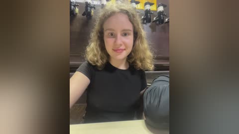Media: Video of a young woman with curly brown hair, wearing a black shirt, sitting at a wooden desk with a microphone and toy figures in the background.