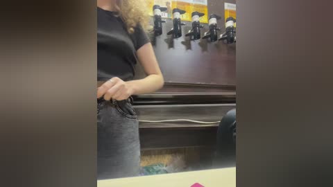 Media: A video of a woman with curly blonde hair adjusting her black jeans in a dimly lit, cluttered room with a wooden table and a wall-mounted microphone setup.