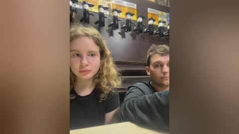 Media: Video of two young people, a blonde girl and a boy, seated at a table in a dimly lit bar. The background features several black and white beer taps.