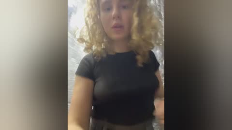 Media: Video of a light-skinned woman with curly blonde hair, wearing a tight black shirt, partially revealing her breasts, standing in front of a blurry background.