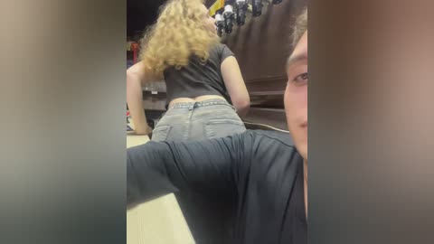 Media: A video of a young woman with curly blonde hair, wearing a black crop top and jeans, bent over a table in a dimly lit room, with another person's face partially visible in the foreground.
