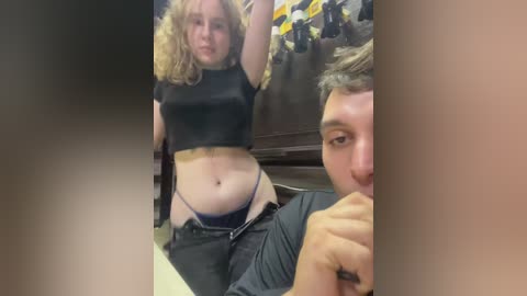 Media: Video of a young woman with curly blonde hair in a black crop top, revealing her midriff, and blue thong, standing provocatively. A young man with short brown hair, wearing a black shirt, sits nearby, looking at her.