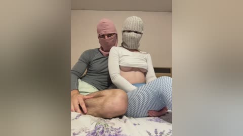 Video of a man in a pink mask and grey shirt, sitting on a bed with a woman wearing a grey knitted mask and blue fishnet tights, in a plain room.