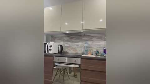 Media: A modern, minimalist kitchen with beige cabinets, a white oven, black kettle, and light gray and white mosaic tile backsplash. The countertop is dark gray, and a small white stool is positioned near the oven.