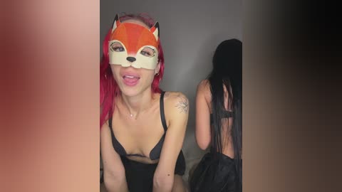 Video of a light-skinned woman with vibrant red hair, wearing a fox mask and black lingerie, standing against a dark wall, with another person partially visible behind her.