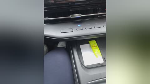 Media: Video of a car interior with a black dashboard featuring a digital display and audio controls, a smartphone charging in the center console, and a person's leg visible in dark blue pants.