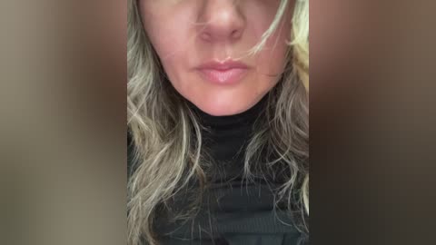 Close-up video of a woman with fair skin, wavy blonde hair, and light pink lips. She's wearing a black turtleneck sweater, partially obscured by blurry brown background.