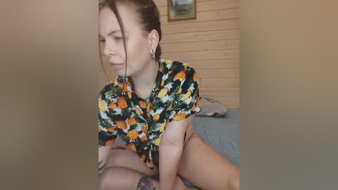 Video of a fair-skinned woman with brown hair in a ponytail, wearing a floral-patterned top, sitting on a bed in a rustic wooden-paneled room with a framed landscape painting on the wall.