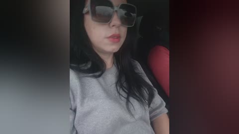Video of a woman with long black hair, wearing oversized square sunglasses, gray T-shirt, and red lipstick, sitting in a car with a red interior.
