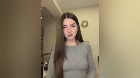 Media: Video of a young woman with long, straight brown hair, fair skin, and a slender physique, wearing a fitted gray top, standing in a dimly lit, sparsely furnished room with beige walls and a clock.
