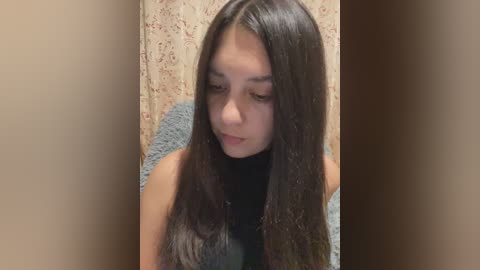 Video of a young woman with long, straight, dark brown hair, wearing a black sleeveless top, looking down thoughtfully. The background features a beige floral-patterned curtain.