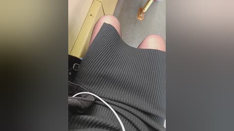 Video of a person's legs wearing a ribbed, black skirt with white trim, seated in a beige chair, with a yellow object, possibly a box, visible in the background.